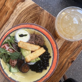 Guava Tree Cafe - A colorful bowl contains salad, shredded beef, plantains, and black beans, with a small container of sauce, on a wooden table with a drink nearby. - Latin American Near Me - Albuquerque, New Mexico