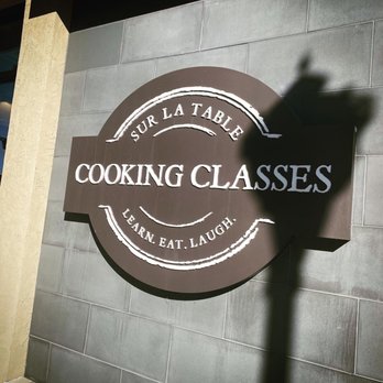 Sur La Table - Sign for the shop and class - A circular sign for Sur La Table displays the text "Cooking Classes" and "Learn. Eat. Laugh." on a gray tiled wall, with a shadow cast across it. - Kitchen Supplies Near Me - Centennial, Colorado