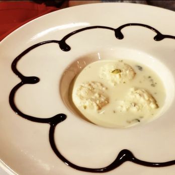 Nirvana Indian Kitchen - Bhel puri - A white ceramic bowl holds an Indian dessert featuring three round dumplings in a milky, slightly thickened sauce. The bowl's rim is decorated with a pattern of chocolate syrup, creating a decorative border. The background shows a reddish surface, possibly a table, with a blurred view of a person or object. The dessert appears to be a traditional Indian sweet, served in a restaurant setting. - Indian Near Me - Allendale, New Jersey