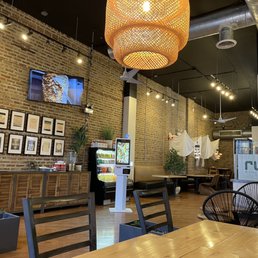 the dining area - The photo depicts the interior of Rumi Middle Eastern Grill in Chicago, with exposed brick walls, a mounted television, framed artwork, and a vending machine. The space includes a mix of seating options such as chairs and booths, with decorative elements like plants and hanging lights. The ceiling has visible ductwork and ceiling fans, creating a contemporary ambiance suitable for casual dining.