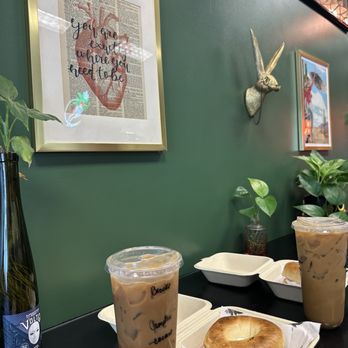 Wild Pine - Campfire iced coffee and bagel with cream cheeese - The image shows two iced coffee drinks in clear plastic cups with handwritten labels, along with two bagels in white takeout containers on a black table. The background features a green wall decorated with framed artwork, including a quote and a deer head sculpture, as well as potted plants. - Coffee & Tea Near Me - Peoria, Arizona