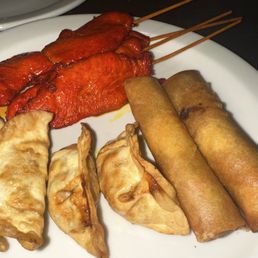Appetizer Sampler platter - A white plate holds Thai appetizers such as satay skewers, spring rolls, and fried dumplings, served with a dark background.