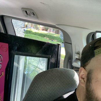 GlassTech - The image shows a person with a beard, wearing sunglasses and a black shirt, seated in the front passenger seat of a vehicle. The person's face is partially visible, with the right side of their face and ear shown. The vehicle interior includes a gray fabric seat and a ceiling with a small light fixture. Behind the person, a large black object, possibly a display or equipment, is positioned, partially blocking the view of the window. The window reveals a blurred outdoor scene with greenery and a building in the background. - Auto Glass Services Near Me - Chico, California