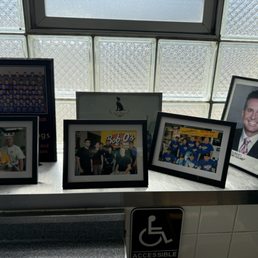 Pictures - Six framed photographs are placed on a windowsill in front of a glass block window. The images include group photos, a portrait of a smiling man in a suit, and pictures of people at a restaurant and a store. The window allows natural light to illuminate the display, and a wheelchair accessible sign is visible below the windowsill.