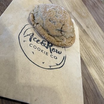 Elevate Coffee - Molasses cookie - A baked cookie dusted with powdered sugar is positioned on a paper bag featuring the Ace & Raw Cookie Co. logo. The cookie appears to be large and slightly cracked, with a textured surface. The paper bag is on a wooden table, and part of the table's surface is visible in the background. The image captures a close-up view of the cookie and branding, emphasizing the bakery's product presentation. - Coffee & Tea Near Me - Phoenix, Arizona