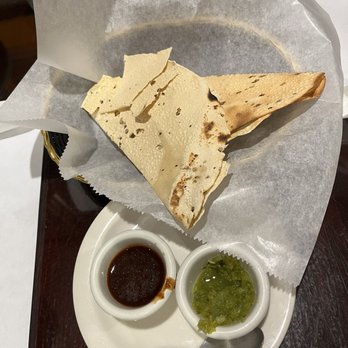 Namaste - Complementary - A white plate holds a serving of crispy papadum, with two small bowls of dipping sauces—one dark soy sauce and one green chutney. A metal spoon rests on the plate, and the plate is placed on a dark wooden table. The papadum is partially covered with a piece of parchment paper. - Indian Near Me - Rio Rancho, New Mexico