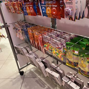 Lick - Some of the 18+ candy - The image shows store shelves stocked with a variety of candies and novelty items. The top row features packages of gummy pecker rings in red, orange, and blue packaging. Below, there are bags of Weenie Chew candies and sour candies with colorful packaging. The display is organized with hooks and racks, and the background reveals a brightly lit retail environment with other store sections visible. - Desserts Near Me - Ledyard, Connecticut