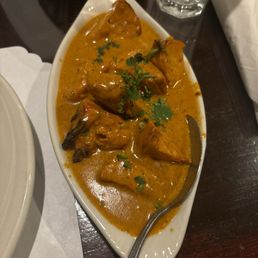 Chicken Tikka Masala - A serving of chicken tikka masala with chunks of chicken in a creamy, orange-colored sauce is shown in an oval white dish. The dish is garnished with chopped cilantro and has a metal spoon resting inside. The setting includes a glass of water and a napkin on a dark wooden table, indicating a restaurant environment.