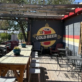 The 377 Brewery - An outdoor seating area at a brewery features wooden tables, metal chairs, potted plants, and string lights, with a mural advertising cold beer. - Brewpubs Near Me - Albuquerque, New Mexico