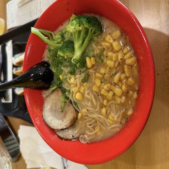Bubblebee X Ramen - R6. Black Garlic Tonkotsu Ramen R6. - A bowl of ramen with a creamy broth contains broccoli florets, yellow corn kernels, and slices of pork. The ramen noodles are submerged in the broth, and a black spoon rests inside the bowl. The bowl is placed on a wooden table with chopsticks, a paper menu, and a glass of water nearby, indicating a dining setting. - Ramen Near Me - Ashburn, Virginia