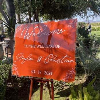 Villa de Amore - A decorative wedding welcome sign is displayed outdoors, featuring a vibrant orange background with floral accents in the corners. The sign reads "Welcome to the wedding of Amylee & Christian" and includes the date "05.19.2023." It is mounted on a wooden easel and surrounded by lush greenery, trees, and plants, with a person visible in the background. The setting appears to be a garden or vineyard venue, suitable for outdoor wedding ceremonies. - Venues & Event Spaces Near Me - Temecula, California