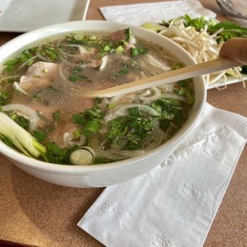 Pho Pasteur 3 - Pho tai - A bowl of Vietnamese pho containing sliced meat, green onions, and herbs, with bean sprouts and vegetables on a side plate. - Vietnamese Near Me - Lancaster, Pennsylvania