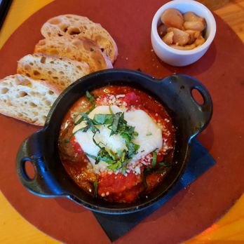 M'tucci's Italian - Meat-a-balls - The image shows a dish of meatballs in tomato sauce, topped with melted cheese and garnished with chopped herbs, served in a small black cast iron skillet. Accompanying the main dish are slices of toasted bread arranged on a round wooden serving board and a small white bowl filled with mushrooms. The setting appears to be a restaurant table, with a wooden surface visible. - Italian Near Me - Albuquerque, New Mexico