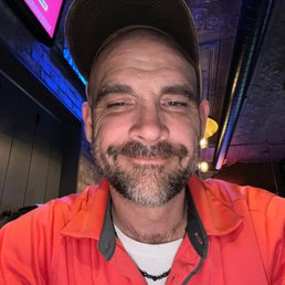 Cant wait to eat some Pizza!!!! - A man with a beard and mustache, wearing a baseball cap and a red Under Armour shirt, takes a selfie in a dimly lit indoor setting.