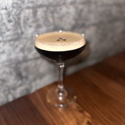 Espresso Martini - A cocktail with dark liquid and a foamy top is presented in a coupe glass on a wooden surface. The drink appears to be a coffee or espresso-based cocktail, with a smooth foam layer and small dark garnishes on top. The background features a gray brick wall, creating a rustic ambiance. The image focuses on the drink, highlighting its presentation and setting in a bar or restaurant environment.