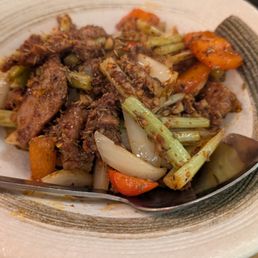 Sizzling Cumin Beef - A ceramic bowl holds cumin beef stir-fry with cooked vegetables including carrots, onions, and celery. The dish appears seasoned with spices, and a metal spoon rests inside the bowl. The background shows a table with papers and a small black dish, indicating a restaurant setting.