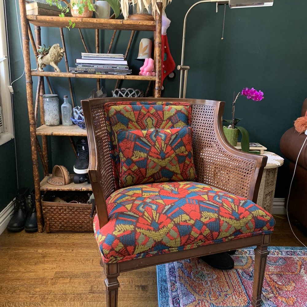 Angelo's Upholstery - Colorfully upholstered antique cane chair, bamboo shelf filled with knickknacks against a dark green wall. - Furniture Reupholstery Near Me - North Hollywood, California Angelo's Upholstery - Colorfully upholstered antique cane chair, bamboo shelf filled with knickknacks against a dark green wall. - Furniture Reupholstery Near Me - North Hollywood, California