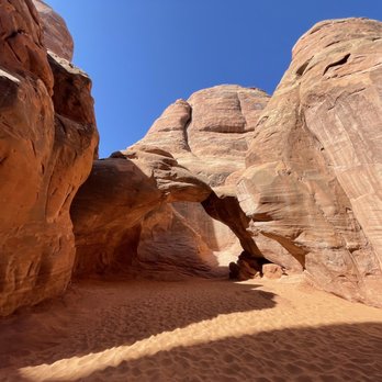 Sand Dune Arch - A sandstone canyon with a natural arch, sandy floor, and towering rock walls, under a bright blue sky, in a desert landscape. - Hiking Near Me - Moab, Utah