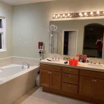 ASR Construction - Original master bathroom, outdated lighting, carpet (?), hulking wood vanity - The image shows a bathroom with a bathtub on the left side, adjacent to a window with frosted glass. The vanity has two sinks, a large mirror above, and a row of lights. Items such as toiletries, a small plant, and a decorative box are on the countertop. A person is visible in the mirror taking a photo, and the bathroom has a neutral color scheme with beige and light green tones. - General Contractors Near Me