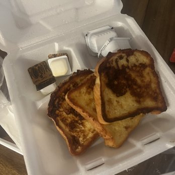 Apollo Beach Diner - French toast - Two slices of French toast with a golden-brown and slightly charred surface are arranged in a white foam takeout container. The container also holds small plastic cups of syrup and cream cheese, indicating a breakfast or brunch meal. The setting appears to be casual, likely from a diner or cafe. - Diners Near Me - Apollo Beach, Florida
