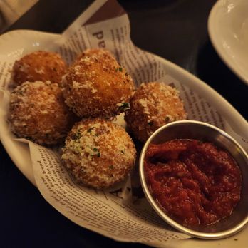 Valentina's - Arancini - A serving of five breaded, fried arancini balls is presented on a dish lined with paper that resembles newspaper print. The arancini are golden brown and coated with breadcrumbs, with visible herbs and seasoning. A small metal cup of marinara sauce accompanies the dish. The background shows other plates and a dark table surface, indicating a restaurant setting. - Italian Near Me - Dublin, Ohio