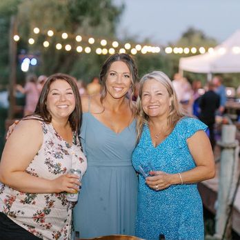 Bronzed Deer studio - Three women stand close together outdoors at an evening gathering near a body of water, with string lights and a tent in the background. - Spray Tanning Near Me - Modesto, California