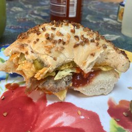 a close up of a sandwich on a plate - A sandwich with various fillings, topped with chopped nuts, on a floral-patterned plate, with a beverage and condiments in the background.