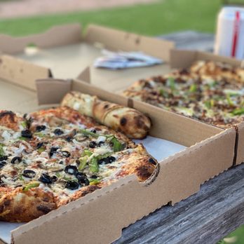 Fired Pizzas - Hulk beast and bbq. - Two slices of pizza in a cardboard pizza box are placed on a weathered wooden table outdoors. The pizza slices have toppings including black olives, green peppers, and melted cheese. A can of soda is visible in the background, along with a blurred outdoor setting with grass and trees. - Food Trucks Near Me - Austin, Texas