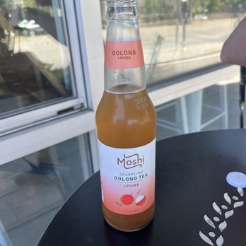 Hashtag Poki - A clear glass bottle of Mosh sparkling Oolong tea with lychee flavor is positioned on a black table. The bottle has a pink and white label indicating the flavor and brand, with a lychee fruit illustration. The background shows a window with a view of trees and a street outside, with natural daylight illuminating the scene. - Poke Near Me - Berkeley, California