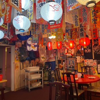 Dragon Sushi - The image shows the interior of a Japanese-themed restaurant with hanging paper lanterns in white and red, decorated with cherry blossom designs. The walls are covered with various Japanese banners, posters, and artwork, creating a vibrant atmosphere. There are tables and chairs arranged for dining, with some tables set with napkins. The decor includes traditional Japanese elements such as a life preserver and anime posters, contributing to an authentic ambiance. - Sushi Bars Near Me - Albuquerque, New Mexico