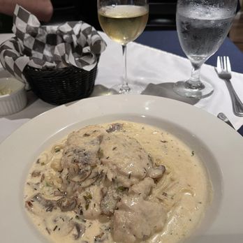Casa Capitano - Pollo Casa Capitano. Delicious! - The image shows a restaurant setting with a white tablecloth, a plate of creamy chicken and mushroom pasta, and two glasses—one with white wine and the other with water. A basket with checkered paper and a small dish are also visible. The background includes chairs and a person wearing a green shirt. The scene suggests a dining experience with Italian-inspired cuisine. - Italian Near Me - Rouses Point, New York