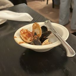 Linguine Alla Pescatore - The image shows a small white bowl filled with leftover shellfish, including dark mussel shells and a piece of crab shell, placed on a dark wooden table. A metal utensil rests on the bowl's edge. The background includes a napkin, a person in gray pants, and a blurred restaurant interior, suggesting a dining setting.