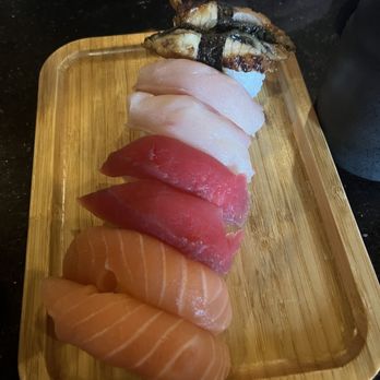 Tsukimi - The image shows a wooden sushi platter with an assortment of sushi pieces arranged in a line. The selection includes slices of salmon, tuna, white fish, eel wrapped with seaweed, and salmon roe. The sushi is placed on a dark surface, with a bowl partially visible at the top and a dark container on the right side. The presentation highlights different types of fish and seafood used in sushi. - Sushi Bars Near Me - Austin, Texas