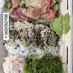 Crab Salad - A takeout container holds a poke bowl with sushi rice sprinkled with black sesame seeds, topped with diced raw fish, vegetables, and garnishes. The bowl includes a portion of wasabi, seaweed salad, and a potato salad. The image appears to be a social media post, showing the food presentation and variety of toppings.
