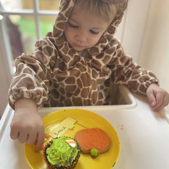 The Classroom - Halloween - A young child wearing a giraffe costume with a hood featuring a giraffe face is seated at a high chair. The child is reaching for cupcakes decorated with green frosting and orange icing, placed on a yellow plate. The background shows a window with a blurred outdoor scene. - Child Care & Day Care Near Me - Vienna, Virginia