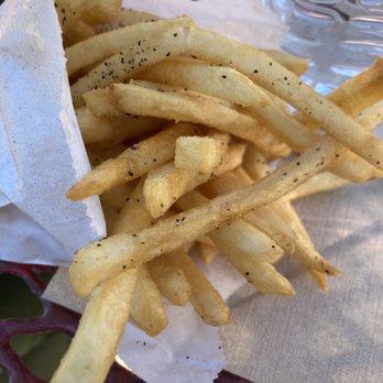 Snarfburger - Fries - French fries seasoned with black pepper are served in a paper-lined basket on a table. The fries are golden and crispy, with visible black pepper seasoning. A glass of water is positioned next to the basket, and the background includes a textured surface and some out-of-focus elements, suggesting an outdoor dining setting. - Burgers Near Me - Boulder, Colorado