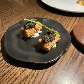 Asador Bastian - Caviar churros - Two pieces of fried food, possibly churros or similar, are placed on a black plate. Each piece is topped with a generous amount of black caviar and garnished with a green chive. The plate is set on a wooden table, with other dishes and utensils visible in the background, indicating a dining setting. - Spanish Near Me - Chicago, Illinois