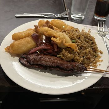 All Seasons Table - COMBO Combo #2 Appetizers Lunch Special - A white round plate contains fried appetizers, including spring rolls and possibly fried bananas, alongside a portion of fried rice with vegetables. A grilled meat skewer rests on the plate. The table has a dark surface with a fork, knife, and napkin, and drinks including water, soda, and possibly iced tea are visible in the background. The setting appears to be a casual Asian fusion restaurant. - Chinese Near Me - Malden, Massachusetts