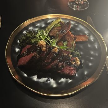 VUE Rooftop - Short rib - A black and gold plate holds sliced cooked steak, broccoli, green beans, potato chips, and garnishes, with a wine glass in the background. - New American Near Me - Washington, DC, District of Columbia