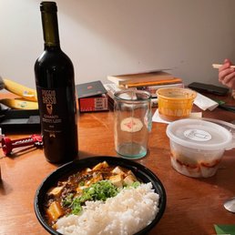 a bowl of rice and a bottle of wine - The image shows a takeout meal served in a black bowl containing white rice, tofu in sauce, and chopped green onions. Surrounding the bowl are a bottle of red wine labeled 'Poggio Basso Chianti,' an empty glass, and several plastic containers with food remnants. The setting appears to be a casual dining or takeout environment with a wooden table and various items in the background.