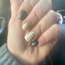 A close-up of a hand with manicured nails, including dark green, gold glitter, and white with gold stripes, held in front of a car interior.