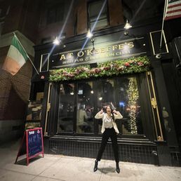Outside - A woman stands on the sidewalk in front of A.T. O'Keeffe's Irish pub in Boston during nighttime, smiling and making a gesture with her hands. The pub's exterior features a black facade with gold lettering, floral decorations, and flags of Ireland and the United States. The scene includes reflections of string lights in the window and a chalkboard sign on the sidewalk.