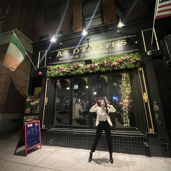 A.T. O'Keeffe's - Outside - A woman stands on the sidewalk in front of A.T. O'Keeffe's Irish pub in Boston during nighttime, smiling and making a gesture with her hands. The pub's exterior features a black facade with gold lettering, floral decorations, and flags of Ireland and the United States. The scene includes reflections of string lights in the window and a chalkboard sign on the sidewalk. - Irish Pub Near Me - Boston, Massachusetts