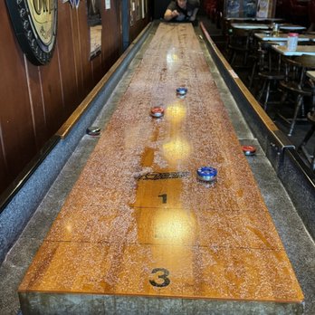 Stober's - A long shuffleboard table with several pucks placed on its surface is featured in a bar interior. The table has numbered sections and a glossy finish. A woman is leaning over the table in the background, and bar decor, including signs and lighting, is visible. The setting includes tables and chairs, with a casual atmosphere. - Bars Near Me - Lansing, Michigan
