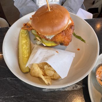 Showroom - Fried chicken sandwich - A fried chicken sandwich featuring a toasted bun, fried chicken fillet, pickles, and a fried egg is presented on a white plate. Accompanying the sandwich are a pickle spear and a few potato chips, with a piece of parchment paper underneath the chips. The plate is on a dark table, and a person wearing a gray jacket is partially visible in the background. - New American Near Me - Frederick, Maryland