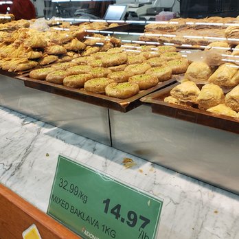 Adonis - A glass display case showcases an assortment of baked goods, including baklava and other pastries, arranged on trays. Price tags indicate the cost of mixed baklava at $14.97 per kilogram and $32.99 per pound. The reflection of the store interior and customers can be seen on the glass surface. The setting appears to be a bakery or grocery store specializing in Middle Eastern or Mediterranean baked items. - Grocery Near Me - Toronto, Ontario