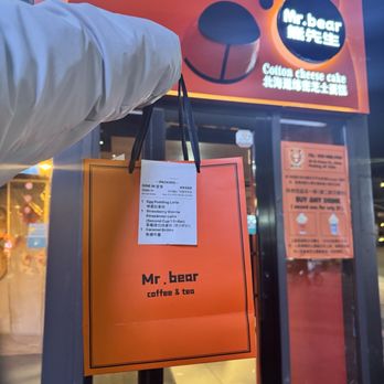 Mr Bear - A person holds a shopping bag outside Mr. Bear coffee and tea shop, which has a bear-themed sign and orange decor. - Desserts Near Me - Queens, New York