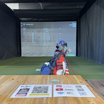 Go Low Golf - An indoor golf simulation room with a large projection screen showing golf range data and statistics. In front of the screen, there is a golf bag with clubs, positioned on a green turf area. The room has dark curtains on the sides and a wooden table with QR codes and informational flyers in the foreground. - Venues & Event Spaces Near Me - Paramus, New Jersey
