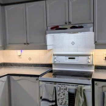 Jdfx Electric - A kitchen features gray upper and lower cabinets, a white stove, and a window above the sink, with under-cabinet lighting. - Electricians Near Me