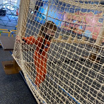 Over the Moon - Climbing in the obstacle course - A child dressed in an orange jumpsuit and wearing a face mask is climbing inside a large rope net structure at an indoor playground. The background shows other play equipment and colorful murals on the walls, indicating a children's activity center with various play zones. - Indoor Playcenter Near Me - Cary, North Carolina